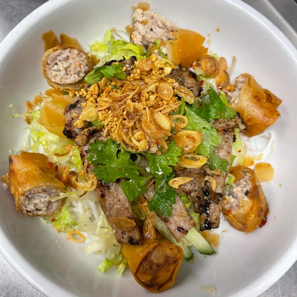 A white bowl filled with vermicelli noodles, grilled pork, crispy spring rolls, and fresh herbs.