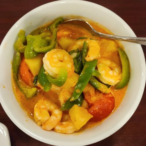 Close-up of Thai red curry with shrimp, green peppers, pineapple, and tomatoes in a white bowl.