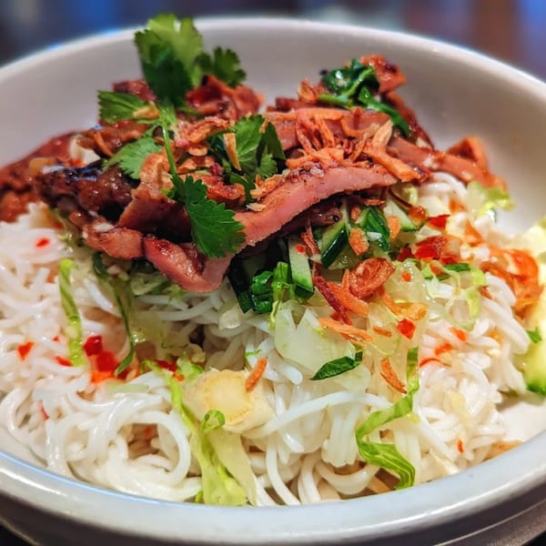 A close-up of a Thai noodle dish with sliced meat, fresh herbs, and crispy fried shallots.
